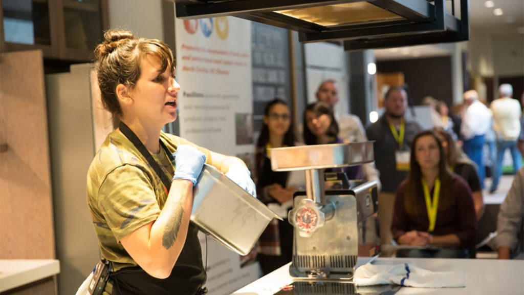 A woman demonstrates meat grinding equipment to an audience